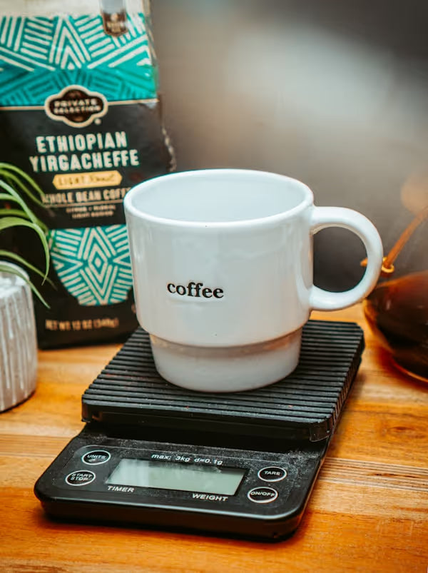 Coffee mug on scale being weighed before brewing. Specialty coffee setup on tabletop with Chemex brewer, coffee beans, and plant. In honor of national coffee day.